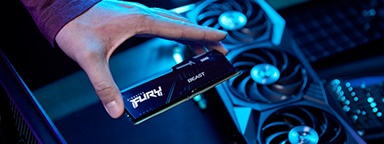 Close-up of a hand holding a Kingston FURY Beast DDR5 RGB memory module, above a desk with PC cooling fans