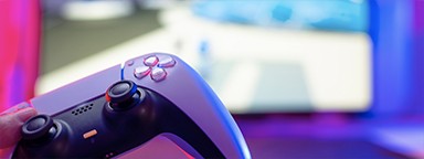 A close-up shot of a PlayStation 5 controller with a blurry background of a game on the TV