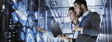 IT specialist holding a laptop talking to a server technician in a server room in a data center with cloud and shield icons on the server racks