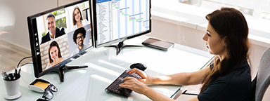 A woman at a desk with two monitors, one with a video meeting showing 4 other people
