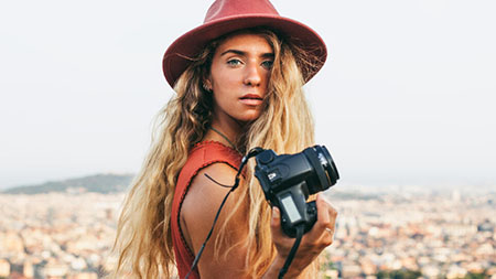 Một cô gái đang đứng trước cảnh thành phố, tay cầm camera và chìa ra cho người xem.