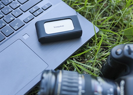 Ổ SSD di động XS2000 Ổ SSD XS2000 trên một chiếc máy tính xách tay trên bãi cỏ, một chiếc camera nằm ở tiền cảnh